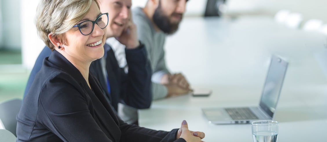 A woman and two men sitting at a conference table with a laptop and a glass of water in front of them.