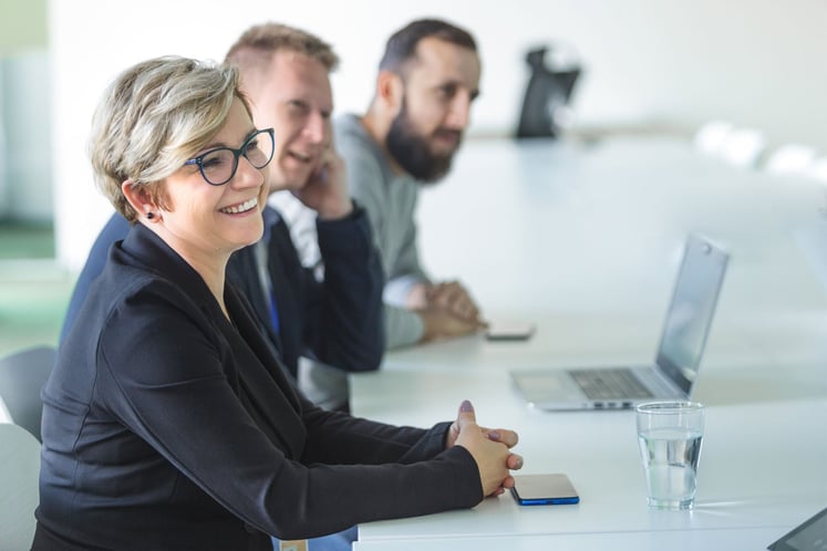 A woman and two men sitting at a conference table with a laptop and a glass of water in front of them. A woman and two men sitting at a conference table with a laptop and a glass of water in front of them.